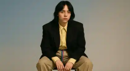 Billie Eilish sitting in a minimalist setting, wearing a black blazer and beige pants