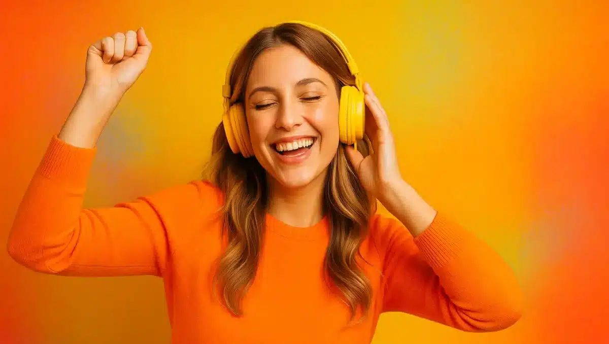 Smiling young woman wearing yellow headphones and orange sweater enjoying upbeat music on a bright orange background.