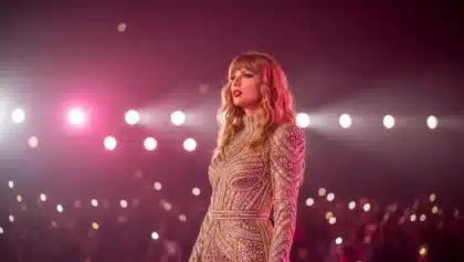 Taylor Swift performing live on stage in a glittering dress, illuminated by concert lights with fans in the background.